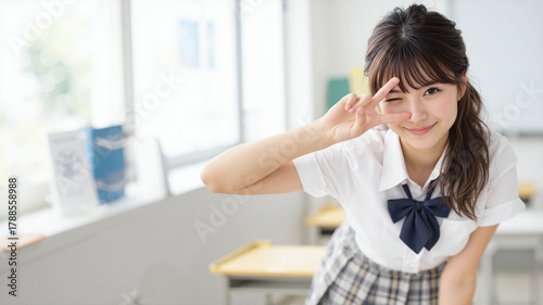 Energetic Asian schoolgirl in uniform gives peace sign and covers eye with playful smirk in bright blurred classroom