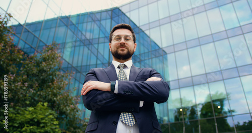 Low-angle close-up of a confident and successful businessman in a suit and tie, looking directly at the camera with crossed arms, standing in front of a modern skyscraper