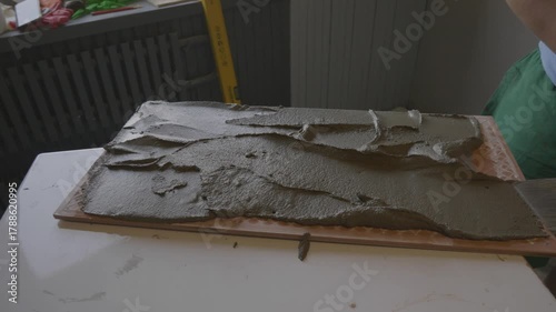 Close-up view of a tiler's hands applying fresh adhesive cement with a putty knife on the back of a ceramic tile before installation, demonstrating a crucial step in home renovation and repair.