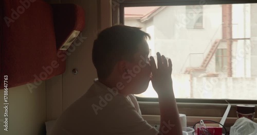 A pensive boy sits in a sleeper train compartment drinking from a glass and watching the scenery pass by the window during a long train ride enjoying the quiet ride The child is traveling on a train.