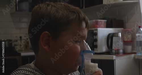 Sick little boy using a nebulizer mask to treat a respiratory illness, breathing in medicated steam to alleviate coughing and congestion while sitting in the kitchen at home.