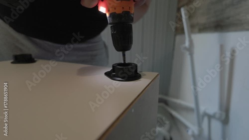 A close-up of a carpenter using a power screwdriver to assemble a new piece of wood furniture. The craftsman carefully tightens a screw into a plastic fitting during a home renovation project.