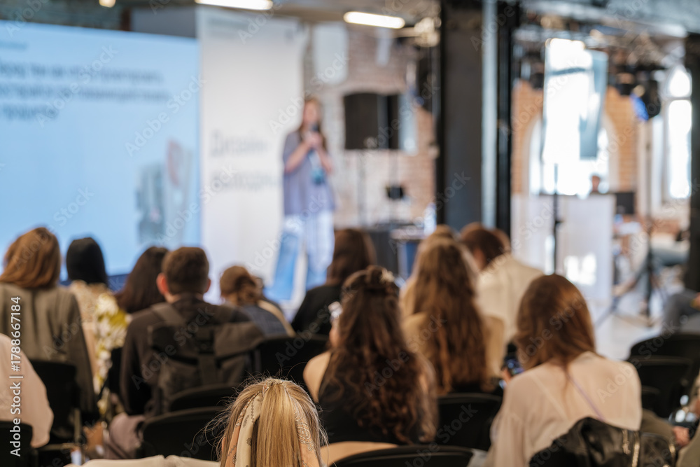 Fototapeta premium Audience attends blurred conference talk with speaker on stage in bright venue.