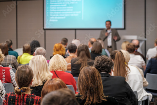 Audience listens to presenter at conference hall with projector screen in background, colorful attendees, business setting, learning atmosphere.