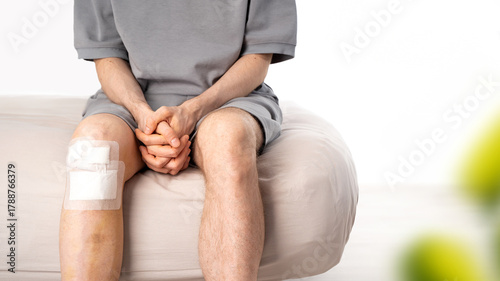 Patient male with bandaged knee sits on bed in light interior. Bruising, medical patch after recent trauma. Concept of injury recovery, first aid, home care, post-surgery healing. Banner. Copy space