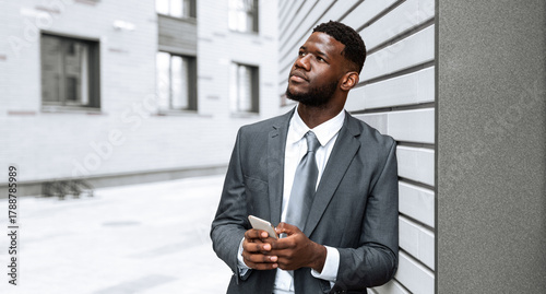 Fotografía A businessman dressed in a suit stands outside a modern building, looking thoughtfully at his smartphone