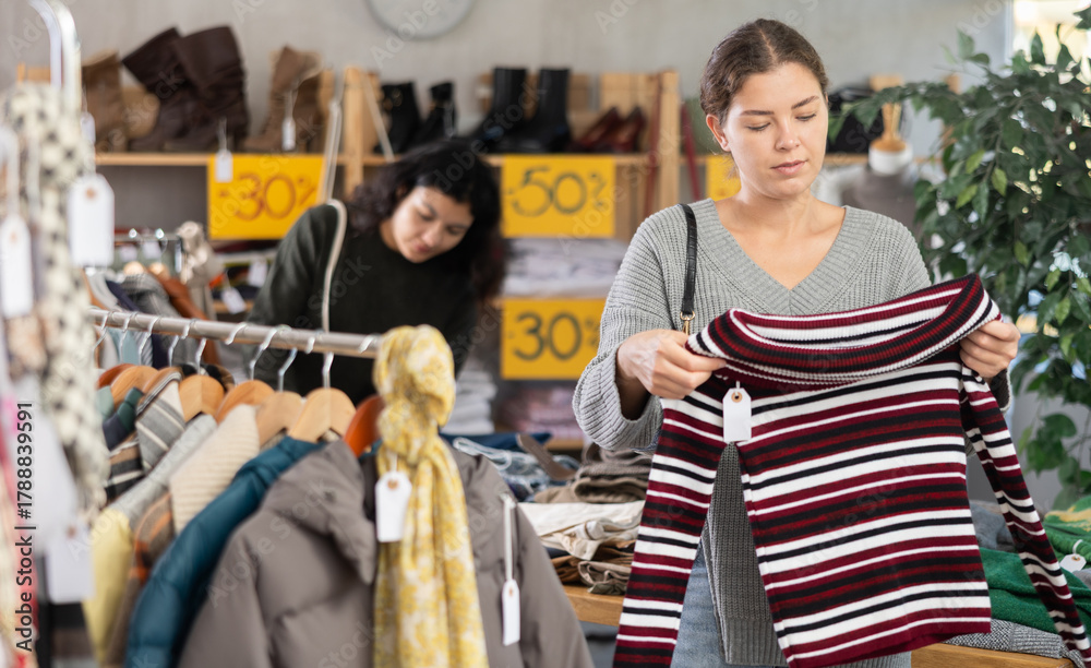 Fototapeta premium European girl is busy looking at clothes items in store, chooses sweater. Young woman enjoys shopping for stylish clothes at clothing store browsing through colorful apparel on display racks