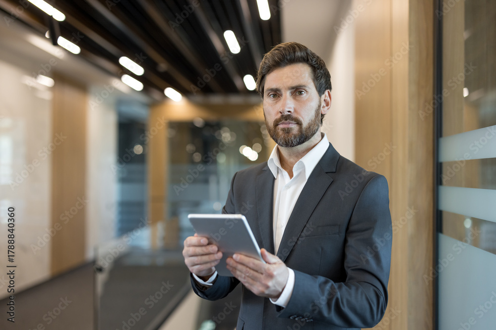 Fototapeta premium Confident bearded businessman standing in a contemporary office hallway, holding a digital tablet, embodying professionalism, technology, and success in a corporate setting