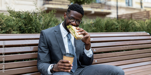 Obraz na plátně A man dressed in a suit sits on a wooden bench in a park, eating a large sandwich with one hand while holding a coffee cup in the other
