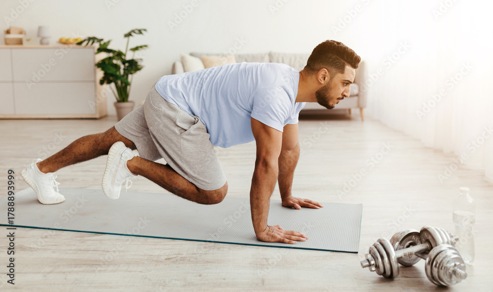 Naklejka premium A man performs a mountain climber exercise on a yoga mat in a bright indoor space. He focuses on his fitness routine while dumbbells rest nearby.
