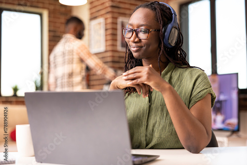 Self employed woman seated with headphones, attentively listening to online webinar on laptop. Black female manager remotely conducting virtual meeting with coworkers, discussing company progress.