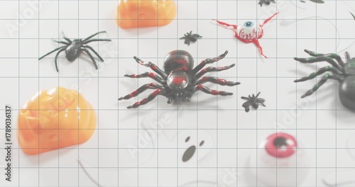 Sitting glossy black-and-red plastic spider on white grid mat at table, with pumpkins and eyeballs