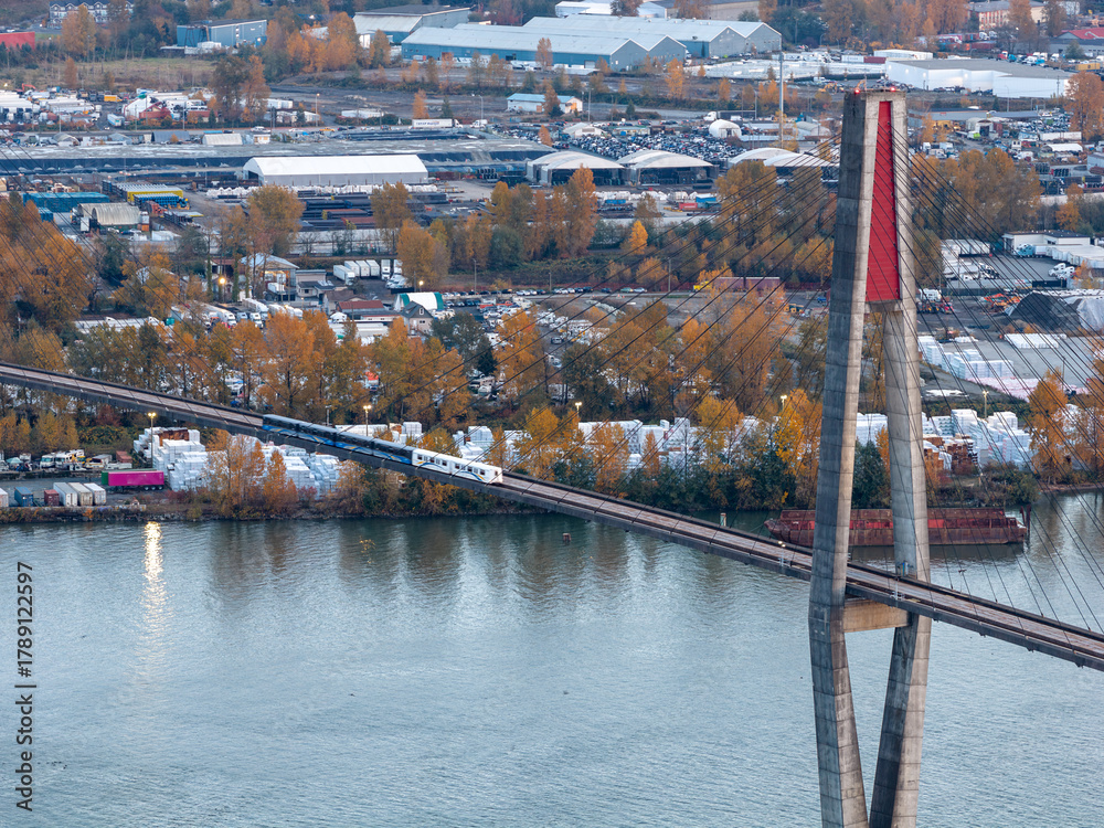 Naklejka premium Suspension Bridge Over River With Train Crossing Amid Industrial Complex and Autumn Trees