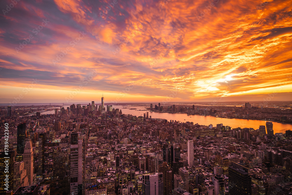 Obraz premium Aerial view of New York City at sunset with vibrant orange sky reflecting in the Hudson River and illuminated skyscrapers under dramatic clouds