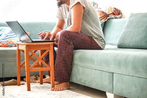 A man in pajamas and glasses, whose faceless is using a laptop. Home office working concept or sunday laziness. Comfortable, friendly atmosphere. Curly hair man work in computer.