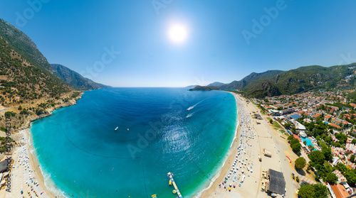 Fototapeta Naklejka Na Ścianę i Meble -  Oludeniz, Turkey. Aerial panorama of vibrant beach under bright summer sun with crowds of sunbathers, turquoise Mediterranean waters and neatly arranged sun loungers. Aerial view.