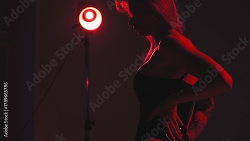Blonde with a beautiful figure in a black patent leather bodysuit poses in a studio. Studio lighting, low exposure, monochrome red light