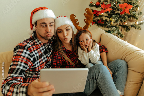 Christmas family wearing Santa hats and reindeer antlers making funny faces while taking a selfie with a tablet, sitting together by a decorated  xmas  tree.