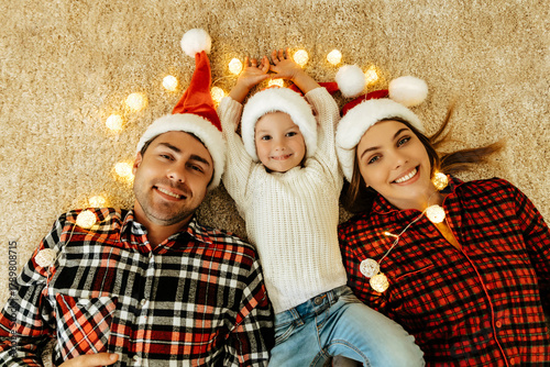 
Dreaming parents and daughter in Santa hats lying on the floor carpet  with glowing lights, sharing love and warmth while celebrating Christmas eve and New Year together.