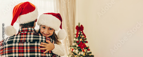 Little girl in Santa hat hugs her father with a joyful smile, both dressed in cozy holiday outfits, with a decorated Christmas tree glowing in the background.
