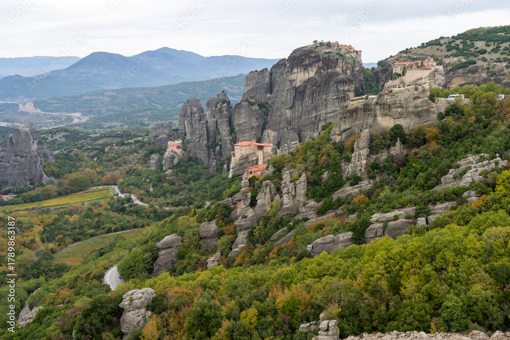 Obraz premium Monasteries crown towering sandstone cliffs in Meteora, surrounded by lush hills and dramatic rock formations.