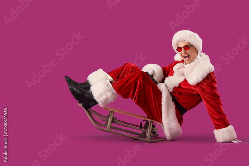 Young Santa Claus sitting on sled against violet background
