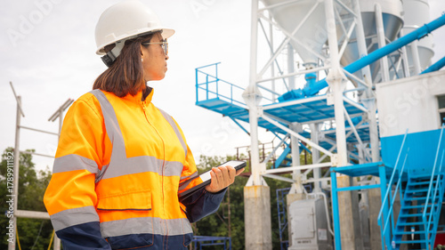 Worker at a construction site wearing high-visibility gear and a hard hat checks equipment with a clipboard in hand. The sun shines on the busy site filled with machinery