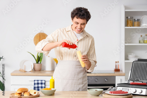 Young man cooking hot dog with ketchup in kitchen