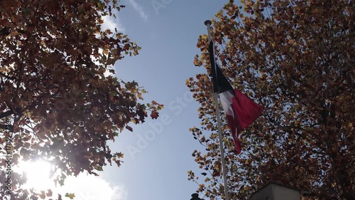 French flag waves proudly atop a stone structure, framed by vibrant autumn trees under a clear blue sky. Golden leaves contrast with tricolor stripes in a striking, patriotic scene.