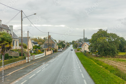 A quiet rural road lined with houses and gardens in a small French village on a cloudy day, showing calm countryside life and traditional architecture.