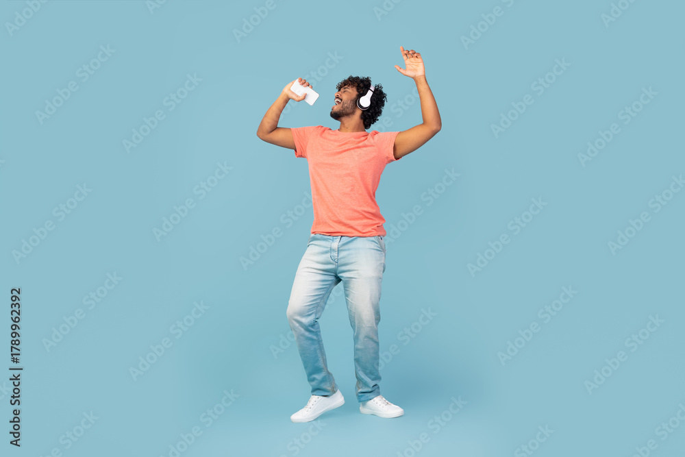 Fototapeta premium An eastern millennial guy with curly hair sings and dances joyfully on a blue studio background. Using a mobile phone as a microphone, he seems to be in a lively moment, enjoying his music.