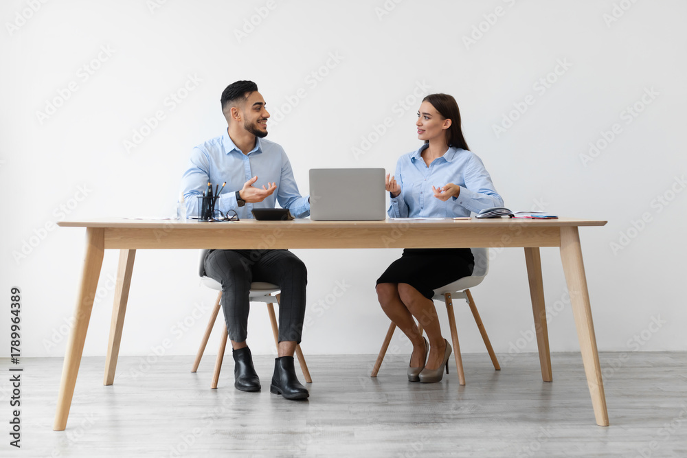 Obraz premium Two colleagues, a smiling woman and a middle eastern man, are seated at a desk with a laptop. They are engaged in a friendly discussion about their business project in a contemporary office.