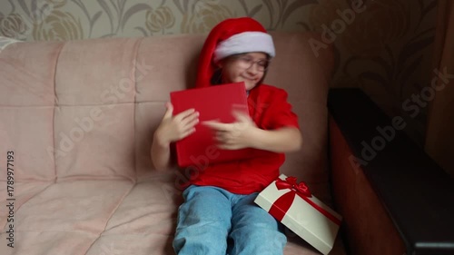 A girl dressed in a red T-shirt and a Santa Claus hat opens a New Year's gift. Christmas. A gift for Christmas