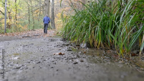 Man in blue waterproof coat walks along a damp muddy footpath in the autumn whilst a drip from above fills a foreground puddle