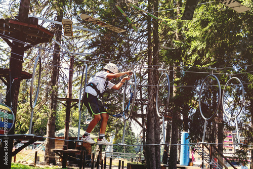 Young adventurer crossing elevated rope course in summer forest canopy challenge park
