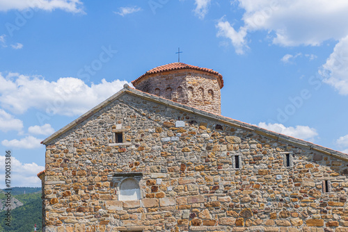 An ancient stone Orthodox church with a red-tiled dome and cross stands under a bright sky