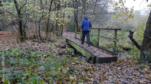 A man walks over a wooden bridge in the Autumn, he's wearing warm outdoor clothing, and the leaves have fallen onto damp ground.