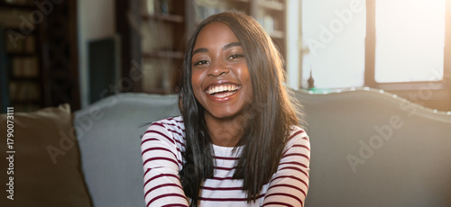 A young woman with long dark hair smiles brightly while sitting on a comfortable couch. Sunlight filters through a window, creating a warm and inviting atmosphere in the cozy room filled with books.