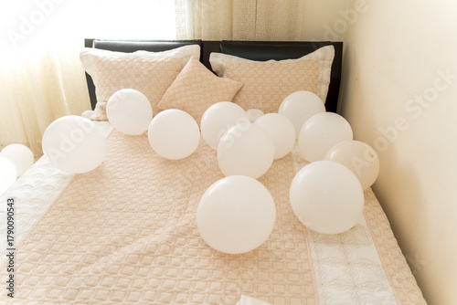 White balloons are spread across a neatly made bed with light-colored bedding. Soft sunlight shines through the window, adding warmth to the scene.