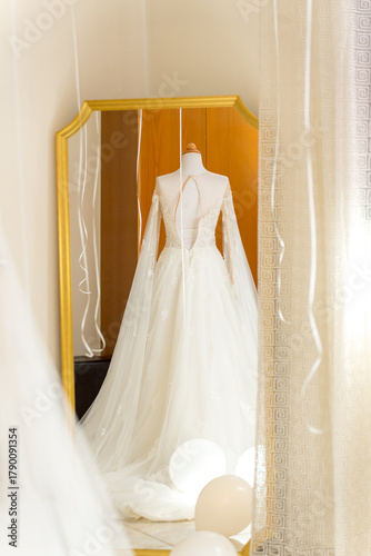 A beautiful bridal gown stands in a fitting room, showcased in front of a full-length mirror. Soft lighting accentuates the gown's lace and flowing fabric against a warm backdrop.