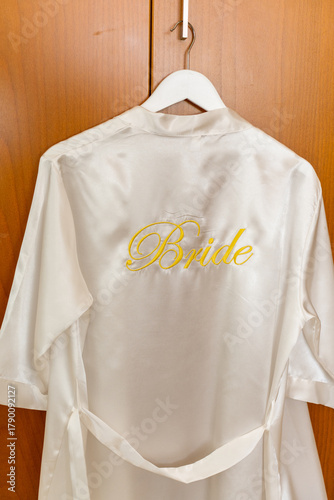 A beautiful satin bridal robe hangs on a wooden closet door. The robe is white with the word 'Bride' elegantly embroidered in yellow. This detail hints at the preparation for a special wedding day.