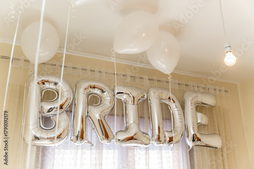 In a cheerful indoor space, silver balloons spelling BRIDE hang from the ceiling, complemented by white balloons. The soft lighting adds warmth to the festive atmosphere.