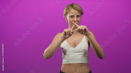 Woman crosses index fingers in magenta studio showing a clear no gesture, frowning and pointing with hands near chest in a white cami top; refusal disapproval warning.
