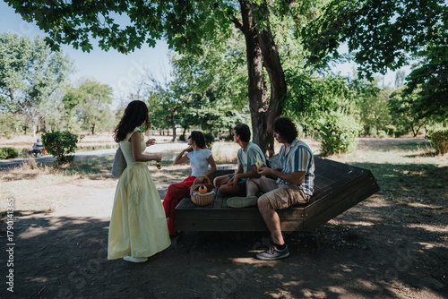 A small group of friends enjoys a casual outdoor gathering in a sunny park. A woman in a yellow dress chats with others on a wooden lounge as a picnic basket sits nearby.