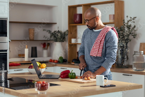 Shaved head man learning to cut vegetables through laptop in kitchen at home