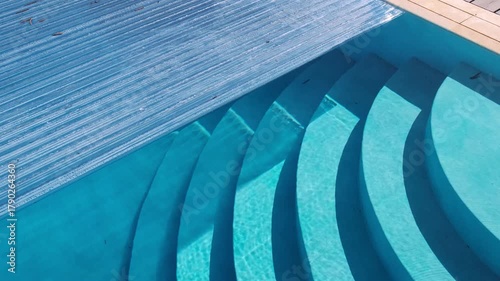 Curved pool steps partially covered by a transparent automatic cover. Bright sunlight and water reflections create a striking geometric pattern
