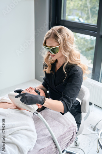 Female cosmetologist receiving laser epilation treatment for man at spa