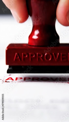 Closeup of a hand holding a red rubber stamp with the word approved embossed on it, ready to stamp a document