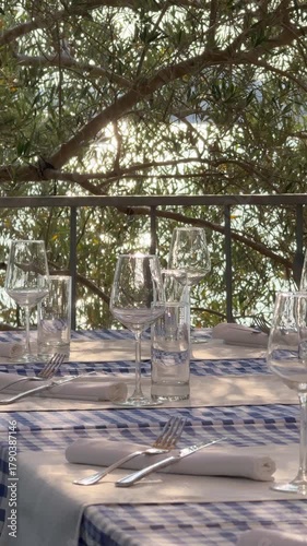 Close up of a beautifully set restaurant table with glassware and plates. In the background, sunlight sparkles on the sea through trees, creating a serene Mediterranean atmosphere.