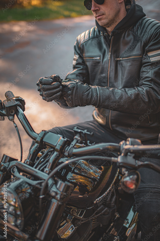 Fototapeta premium Close-up view of a biker in a leather jacket standing near a black custom motorcycle on an autumn road. Concept of freedom, travel and motorcycling lifestyle.